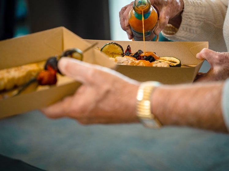 Image of A person pours sauce over grilled vegetables and skewers in a cardboard food tray, whilst another hand holds the tray. Both individuals wear light-coloured clothing.
