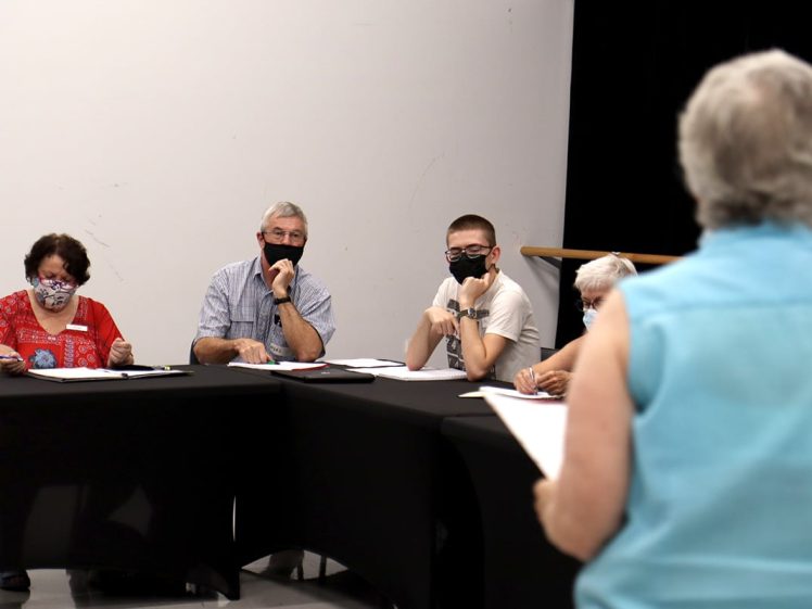 Image of A group of four people wearing masks sit at a table, attentively listening to a standing woman facing them. Papers and pens are on the table as they interact in a meeting or discussion in a bright room.