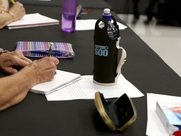 Image of A person writes in a notebook at a table with a striped pencil case, a Hydro 600 water bottle, a purple water bottle, and some papers and items scattered nearby.