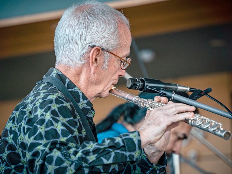 Image of An older man with grey hair and glasses plays the flute into a microphone. He is wearing a patterned shirt and performing indoors. The background is blurred, highlighting the musician.
