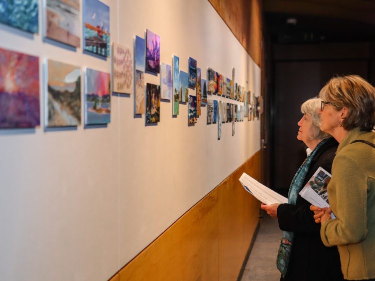 Image of Two women closely view and discuss a variety of colourful paintings mounted on a long gallery wall; each holds exhibition brochures in hand.