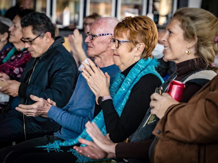 Image of A group of adults sit closely together in a row, applauding and watching something off-camera. They appear engaged and cheerful; one woman wears a bright blue scarf, and another holds a red tin.