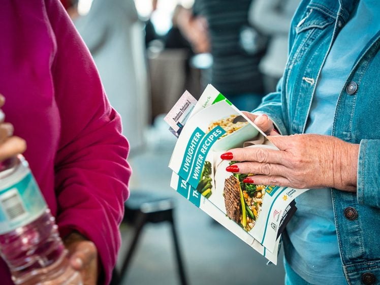 Image of Two people are talking; one holds a water bottle, and the other holds several recipe booklets titled Unlock New Winter Recipes. Both wear casual clothing, and the background shows more people out of focus.