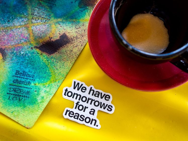 Image of A coffee cup on a red saucer sits on a yellow surface next to a colourful textured mat and a sticker that reads, We have tomorrows for a reason.