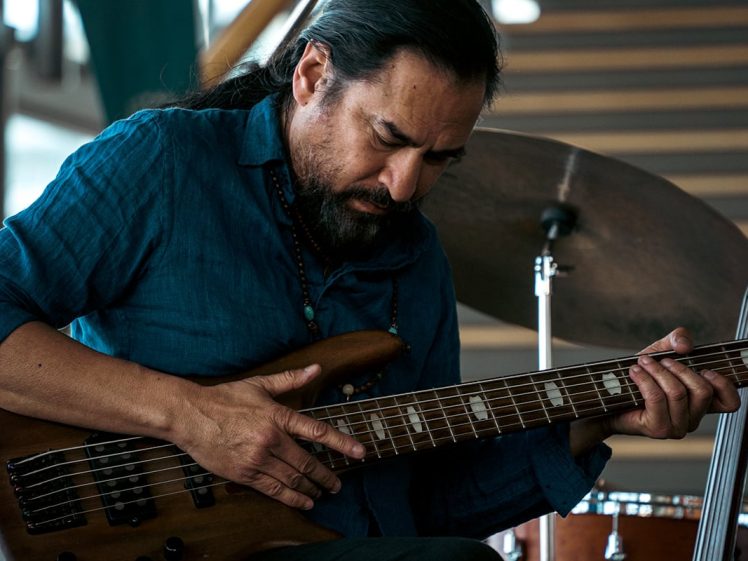 Image of A man with long dark hair and a beard plays a brown electric bass guitar, concentrating intently. He wears a blue shirt and sits indoors, with a drum kit and blurred stairs in the background.