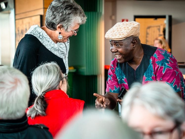Image of A man in a patterned shirt and hat smiles and gestures while speaking to two women in a group. One woman stands and the other sits with her back to the camera. Other people are visible in the foreground.
