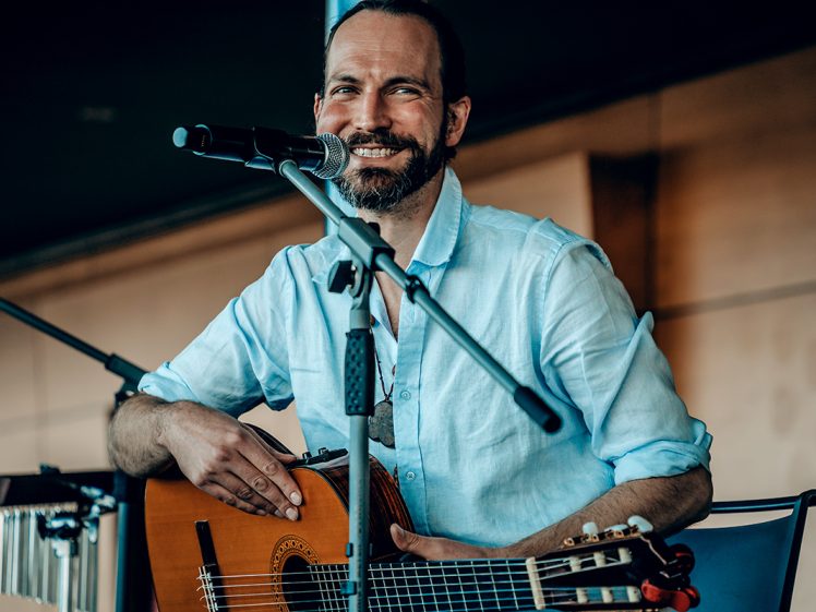 Image of A smiling man with a beard sits and holds a guitar at a microphone, wearing a light blue shirt, on a stage with warm lighting.