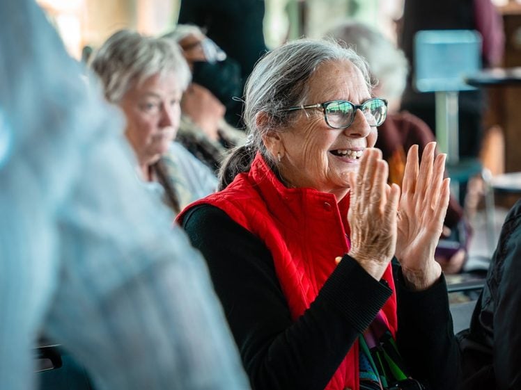 Image of An older woman with grey hair, glasses, and a bright red waistcoat sits indoors, smiling and clapping. Other people are visible in the background, creating a lively, social atmosphere.