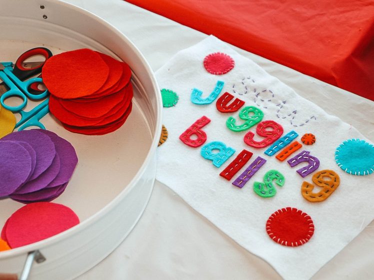Image of A craft table with colourful felt circles, scissors, and a white cloth decorated with the words “juggling balls” in bright, stitched felt letters, surrounded by felt patches.