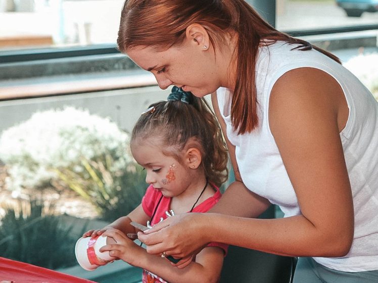 Image of A woman helps a young girl decorate a white cup with stickers and felt-tip pens at a table covered in craft supplies. The girl has face paint and wears a pink top, while the woman is wearing a sleeveless white top.