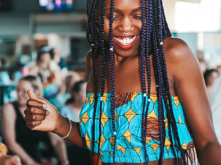 Image of A woman with long plaited hair and a bright, patterned outfit smiles and dances joyfully in a lively indoor setting with a blurred crowd in the background.