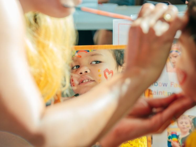Image of A child with face paint looks into a mirror whilst an adult paints a rainbow on their forehead. The child's face is colourful and joyful, with focus on their reflection and the painting process.