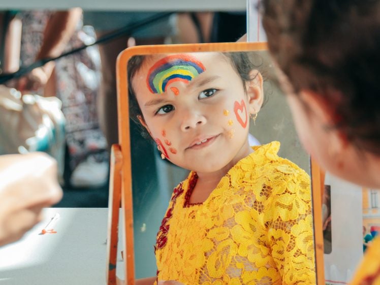 Image of A young child in a yellow lace top looks at her reflection in a mirror. Her face is painted with a rainbow, hearts, and dots, whilst someone’s hand holding a paintbrush is visible in the foreground.
