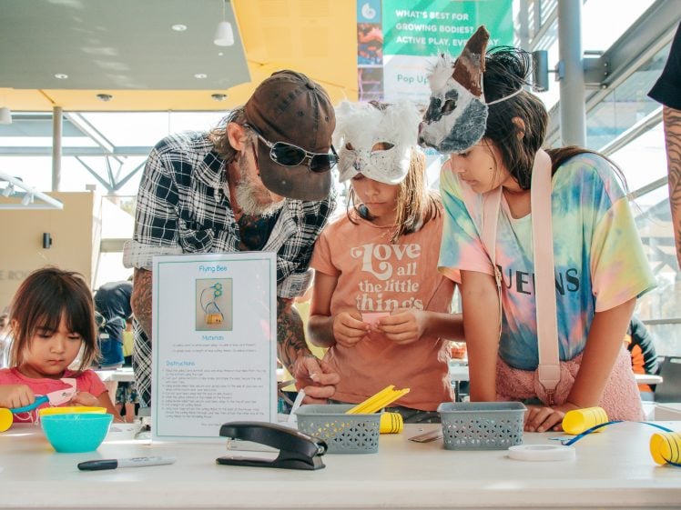 Image of A group of children and an adult, some wearing animal masks, gather around a table to work on a craft project with yellow cups and art supplies in a bright indoor space. A young child concentrates on crafting nearby.