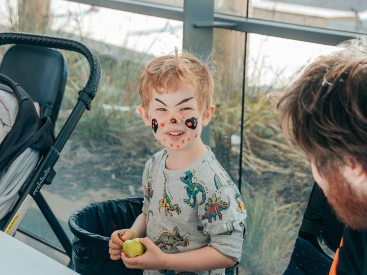 Image of A young child with painted face and a playful expression holds a green apple while standing indoors next to a pushchair and rubbish bin. An adult with a beard is seated nearby, engaging with the child.