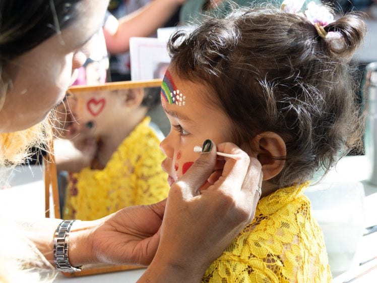 Image of A young child with face paint sits still as an adult adds more designs to her face. The child wears a yellow lace dress and a rainbow with white dots is painted on her forehead. A mirror and painting supplies are nearby.