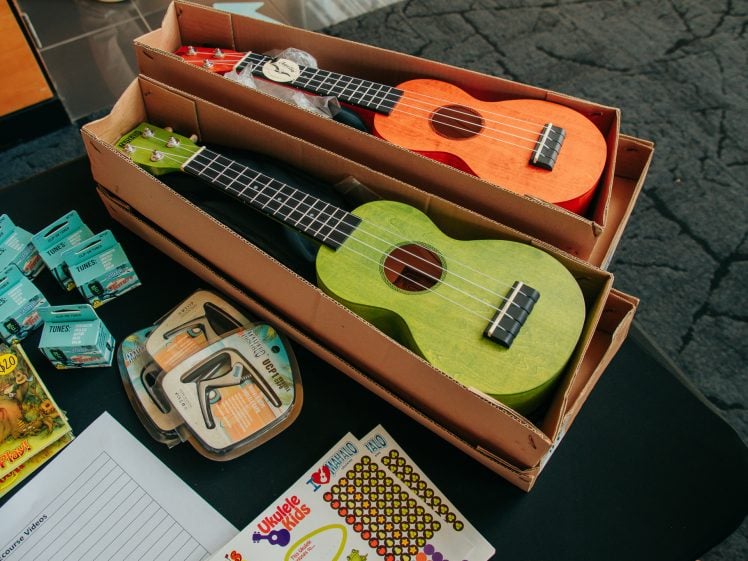 Image of Two colourful ukuleles, one green and one orange, are placed in cardboard boxes on a black table. Nearby are sticker sheets, a triangle instrument, small boxes, and a paper with a sign-up sheet.