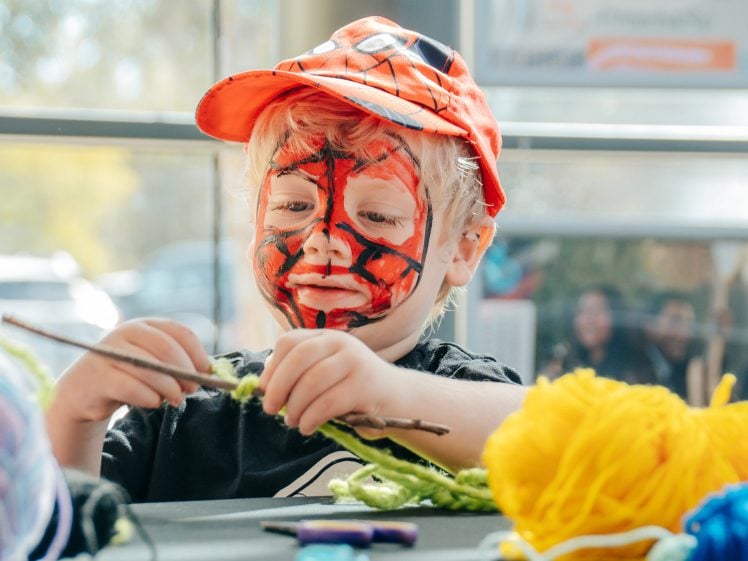 Image of A young child with a Spiderman face paint and red cap is sitting at a table, crafting with colourful yarn and sticks. Sunlight streams in through the window behind him.