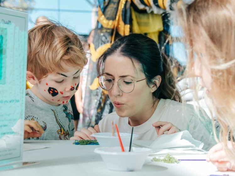 Image of A woman and two children, one with painted spots on his face, are sitting at a table doing an arts and crafts activity. The woman is blowing on a project while the children watch closely. Art supplies are scattered on the table.