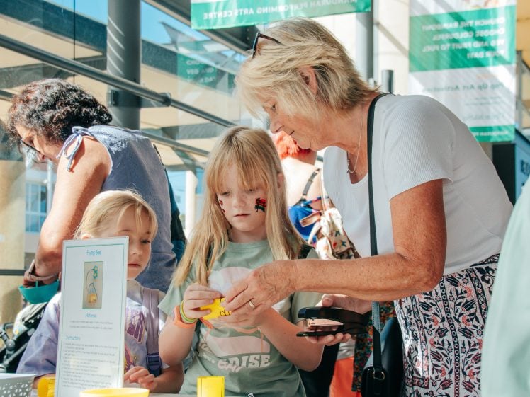 Image of An older woman helps two young girls with a craft activity at a table. One girl has face paint and is focused on assembling something yellow. The setting appears bright and busy, with others visible in the background.