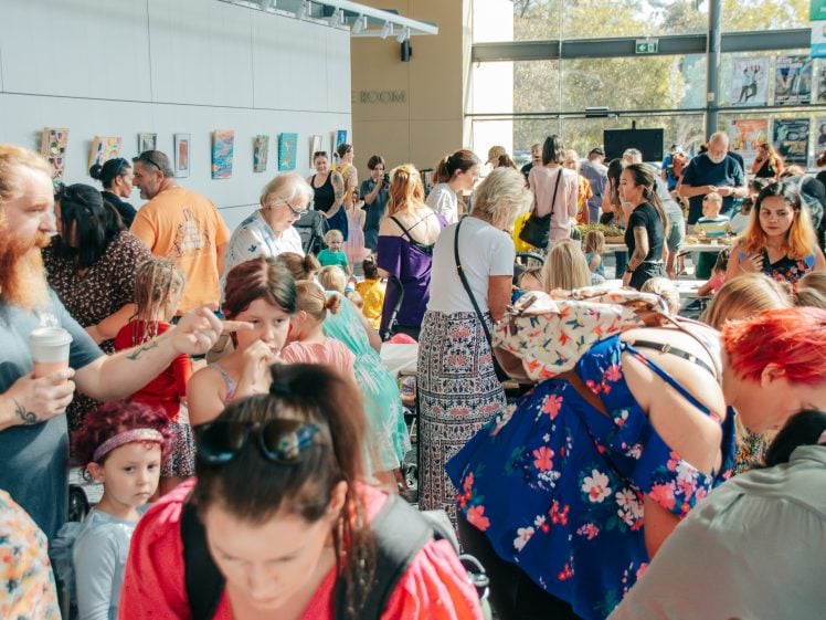 Image of A large group of people, including adults and children, gather indoors at a lively event with tables, artwork on the walls, and sunlight streaming through large windows. Some people are talking, browsing, or holding drinks.