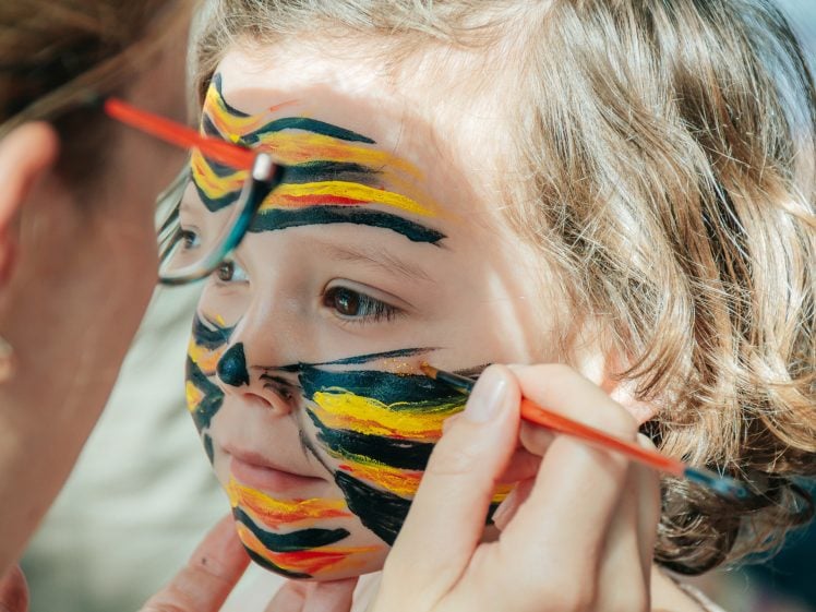 Image of A child with curly hair is having their face painted as a tiger, featuring black, yellow, and orange stripes, whilst an adult carefully applies the paint with a brush.