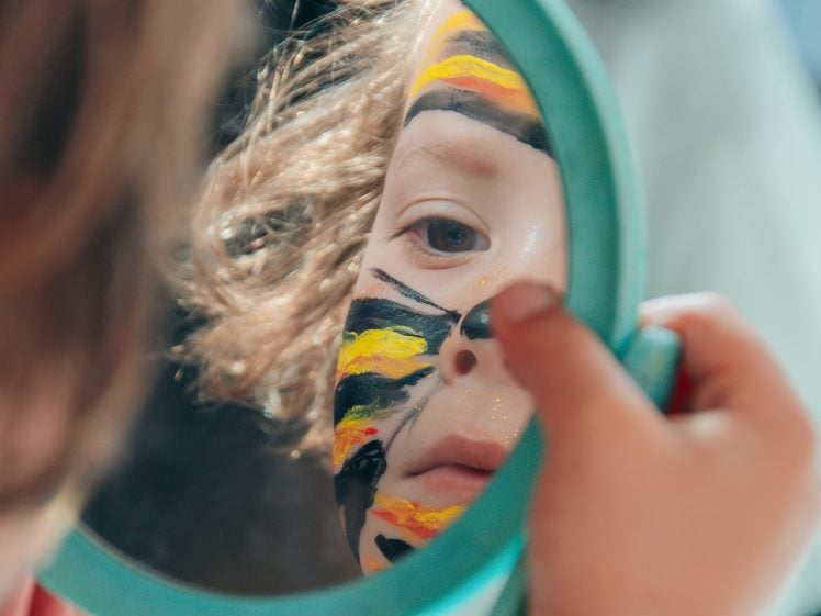 Image of A child with curly hair looks closely into a round mirror, examining their face painted with black, yellow, and orange stripes resembling a tiger.