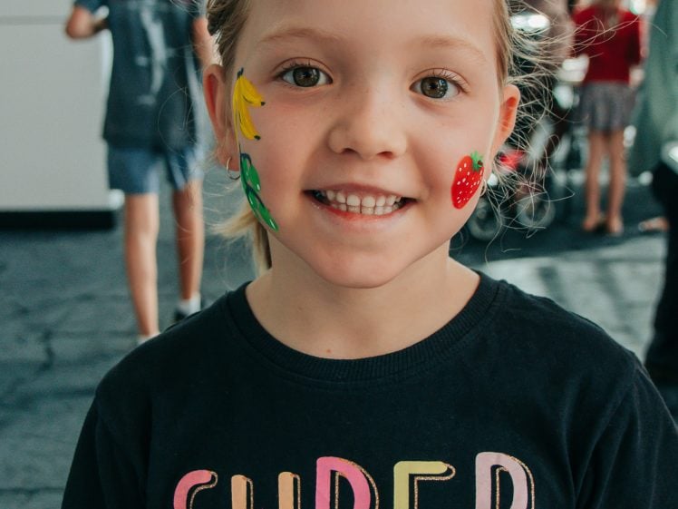 Image of A young child with face paint of a banana and strawberry on their cheeks smiles at the camera, wearing a black T-shirt that says SUPER GOOD in colourful letters. Other people are visible in the blurred background.
