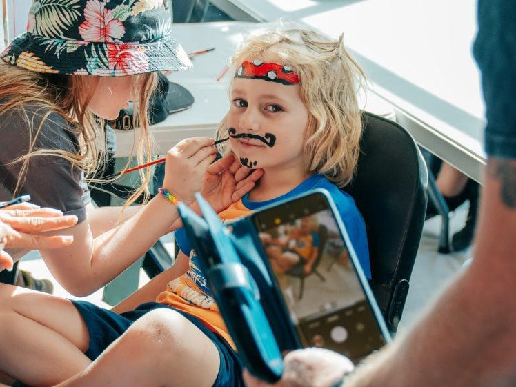 Image of A child sits in a chair getting their face painted with a moustache and beard by another child. The child being painted wears a red headband and blue shirt, while someone holds a tablet, capturing the moment.