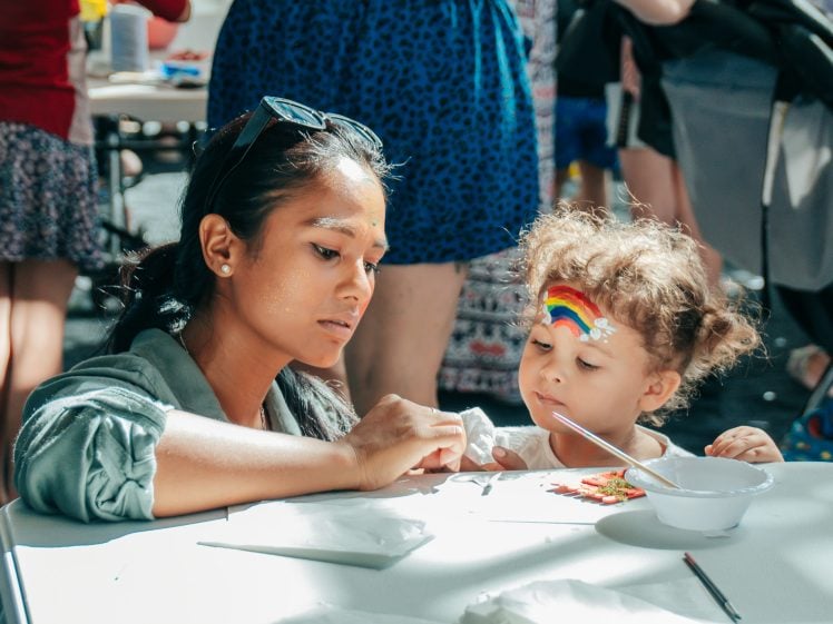 Image of An adult helps a young child with a painted rainbow on her forehead during an arts and crafts activity at a table. Sunlight shines on them, and craft supplies and people are visible in the background.