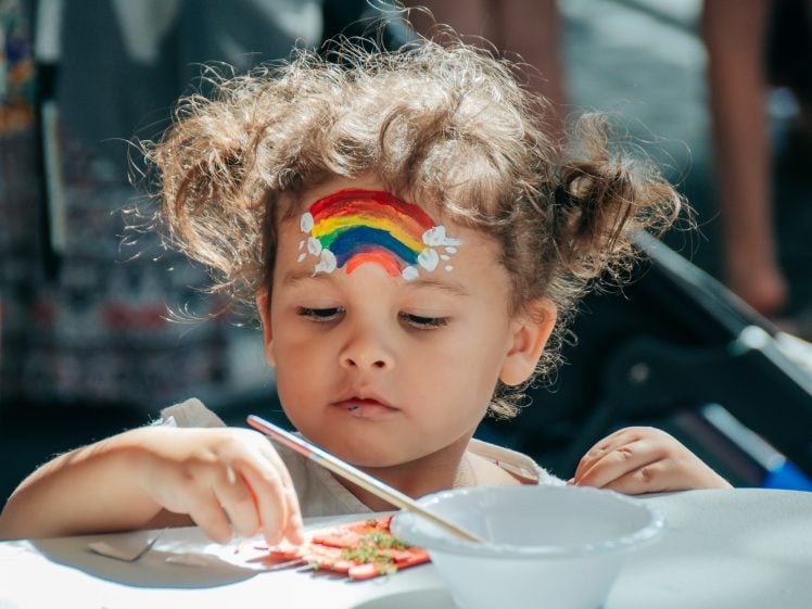 Image of A young child with curly hair and a rainbow painted on their forehead sits at a table, focused on a craft activity, with a white bowl and art supplies in front of them.