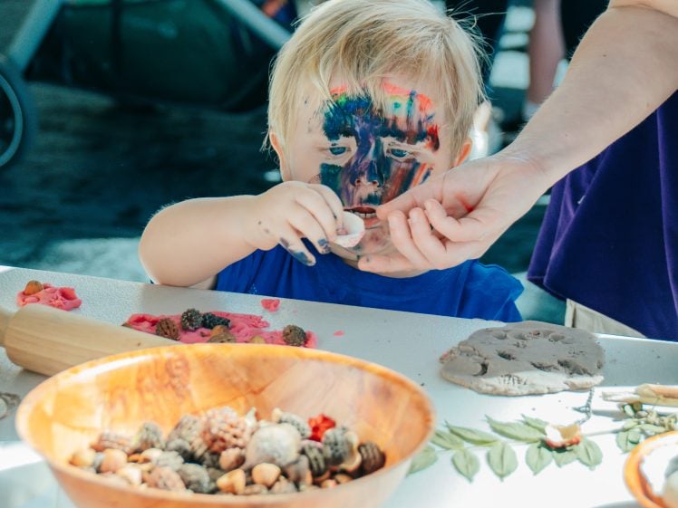 Image of A young child with colourful face paint plays with clay and natural materials at a table, whilst an adult helps guide their hand. Various craft items are scattered on the table.