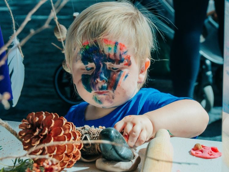 Image of A young child with colourful paint smeared on their face looks down whilst playing with pinecones, stones, and clay on a table. The child wears a blue shirt and is focussed on their activity.