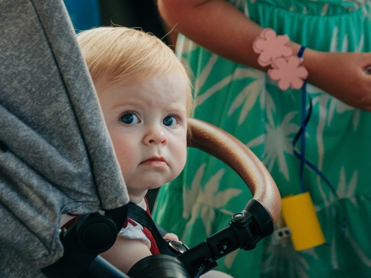 Image of A baby with blond hair sits in a pushchair, looking up with a curious expression. An adult in a green dress with a pink flower bracelet stands nearby, partly out of focus in the background.