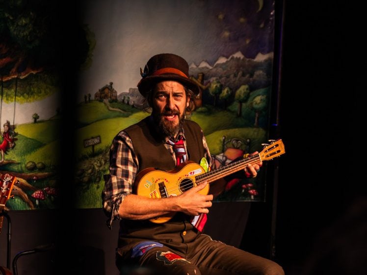 Image of A man wearing a brown hat, checked shirt, waistcoat, and colourful tie plays a small guitar on stage. A whimsical, painted backdrop with trees, mountains, and a starry sky is behind him.