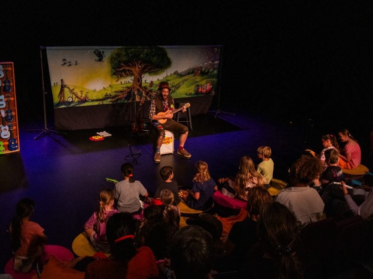 Image of A musician plays a ukulele on stage for an audience of children and adults. The stage is decorated with colourful ukuleles and a backdrop featuring a large tree and bright outdoor scene. The audience sits on cushions and benches.