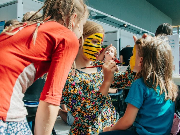 Image of Three children are indoors; one with yellow and black tiger face paint is painting another child's face yellow, whilst a third child watches closely. Sunlight streams through windows in the background.