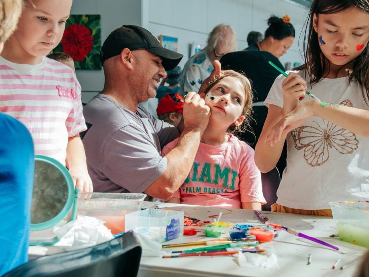 Image of Adults and children sit at a table, painting and doing arts and crafts. A man paints a girl’s face, while another girl paints her own arm. Art supplies and colourful paints are scattered across the table.