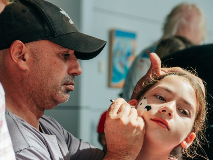 Image of A man wearing a black cap carefully paints a black and white animal face design on a young girl's cheek as she looks off to the side. Other people are blurred in the background.