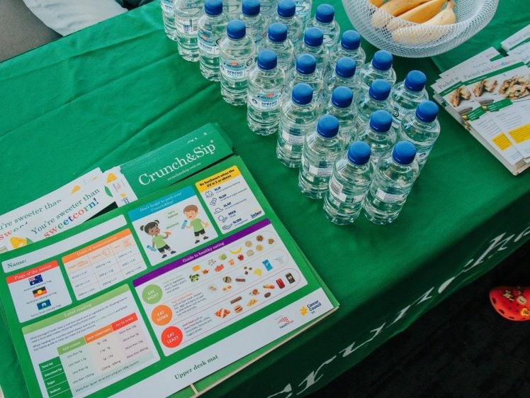 Image of A green table displays bottled water, brochures, and leaflets about the Crunch&Sip healthy eating programme. A basket with bananas is also visible, along with someone wearing red shoes standing nearby.