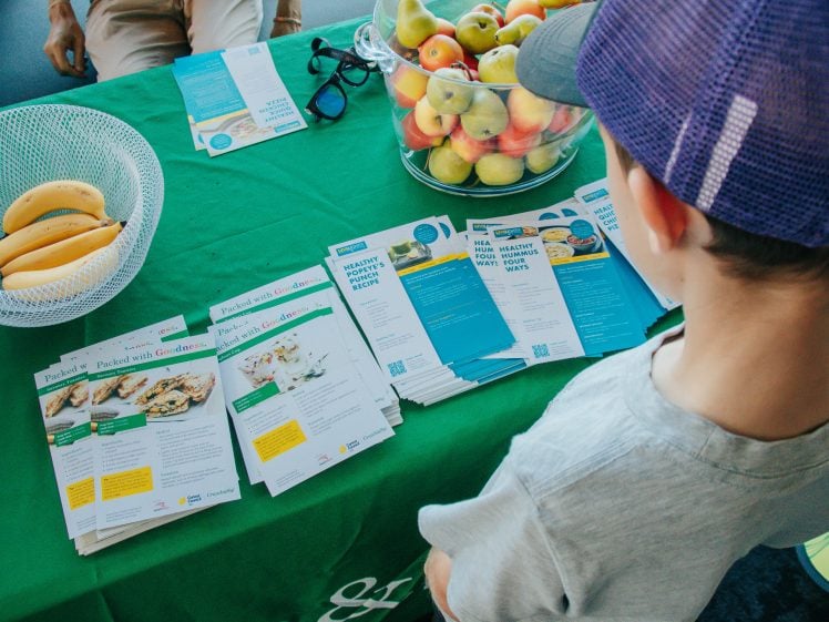 Image of A child wearing a purple cap looks at a table with healthy recipe leaflets, bananas in a white basket, and a bowl of apples. The tablecloth is green and several leaflets display meal ideas and nutrition tips.