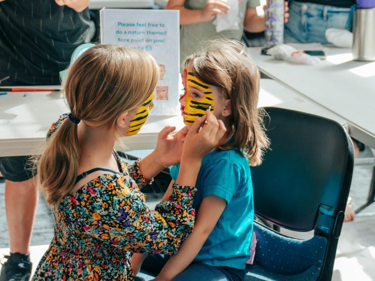 Image of Two children sit together at a table; one is painting the other's face with yellow and black tiger stripes. Other people and art supplies are visible in the background.