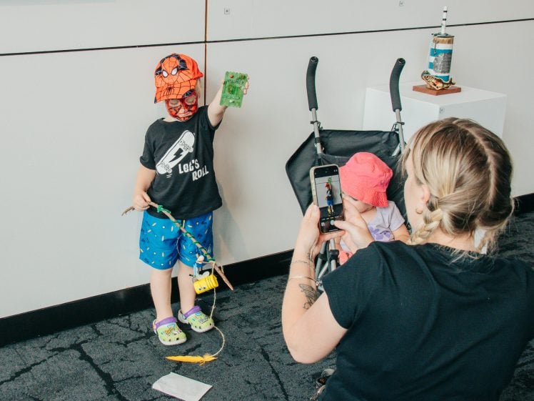 Image of A woman takes a photo of a child in a Spider-Man hat and mask holding up a green object, whilst another child in a pushchair looks down, partially covered by a pink hat.