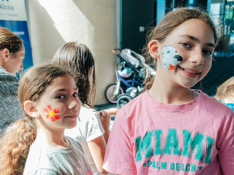 Image of Two girls smiling indoors, both with face paint—one has a red and yellow flower design, and the other has a white dog painted on her cheek. Other children and adults are visible in the background.