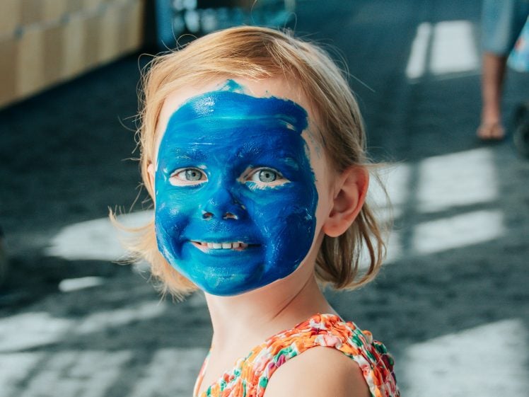 Image of A young child with fair hair smiles at the camera, their entire face covered in bright blue face paint. The child wears a colourful, patterned sleeveless top and is indoors with soft sunlight coming through windows.
