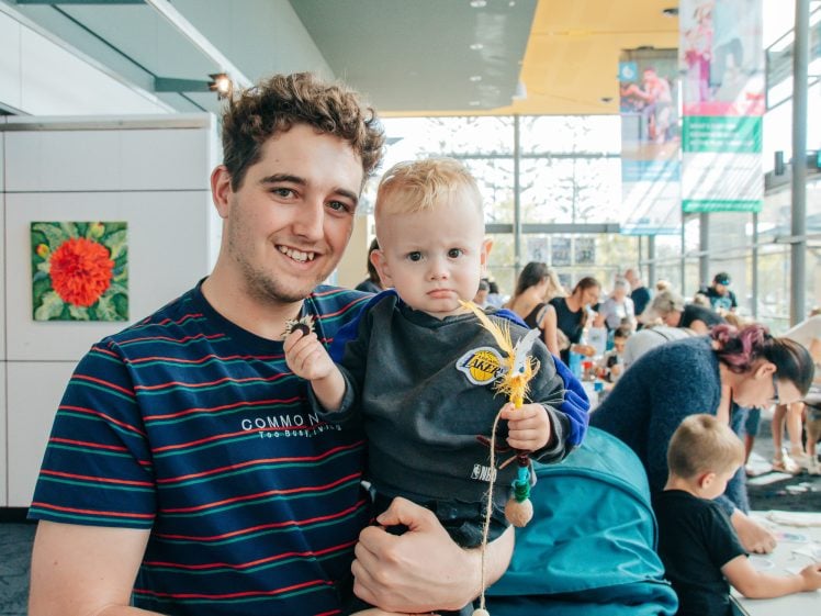 Image of A smiling man holds a toddler indoors at a busy event with people and children in the background. The toddler looks serious and is holding a small craft item. Bright, modern space with art on the walls and large windows.