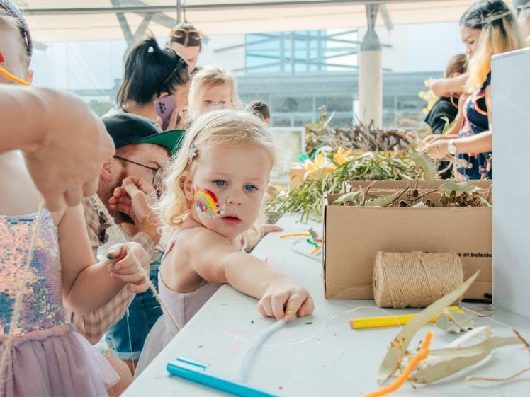 Image of A young girl with a rainbow painted on her cheek reaches for craft supplies at a busy table, surrounded by other children and adults engaged in creative activities.