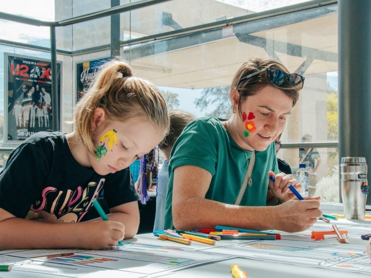 Image of A young girl and an adult with face paint draw with colourful markers at a sunlit table indoors. Art supplies are scattered on the table, and posters are visible on the wall behind them.