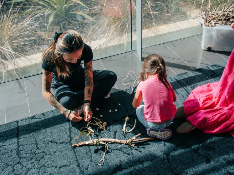 Image of A woman and a young girl sit on a black rug by a large window, making crafts with string, sticks, and shells. Another person in a pink dress is partially visible to the right. Sunlight streams in.