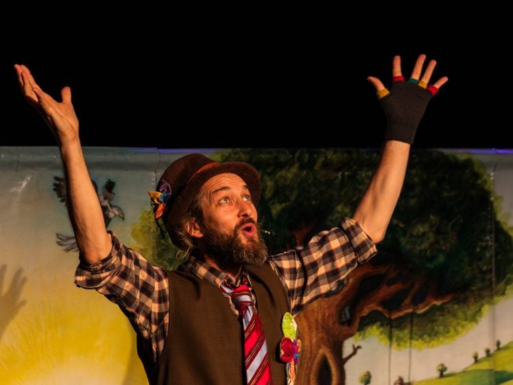 Image of A man with a beard, wearing a hat, checked shirt, tie, and fingerless gloves, stands with arms raised and mouth open in excitement, performing in front of a colourful backdrop with a tree and a sunrise.